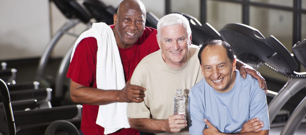 Multi-ethnic senior men (60s) at health club, standing in front of treadmills.